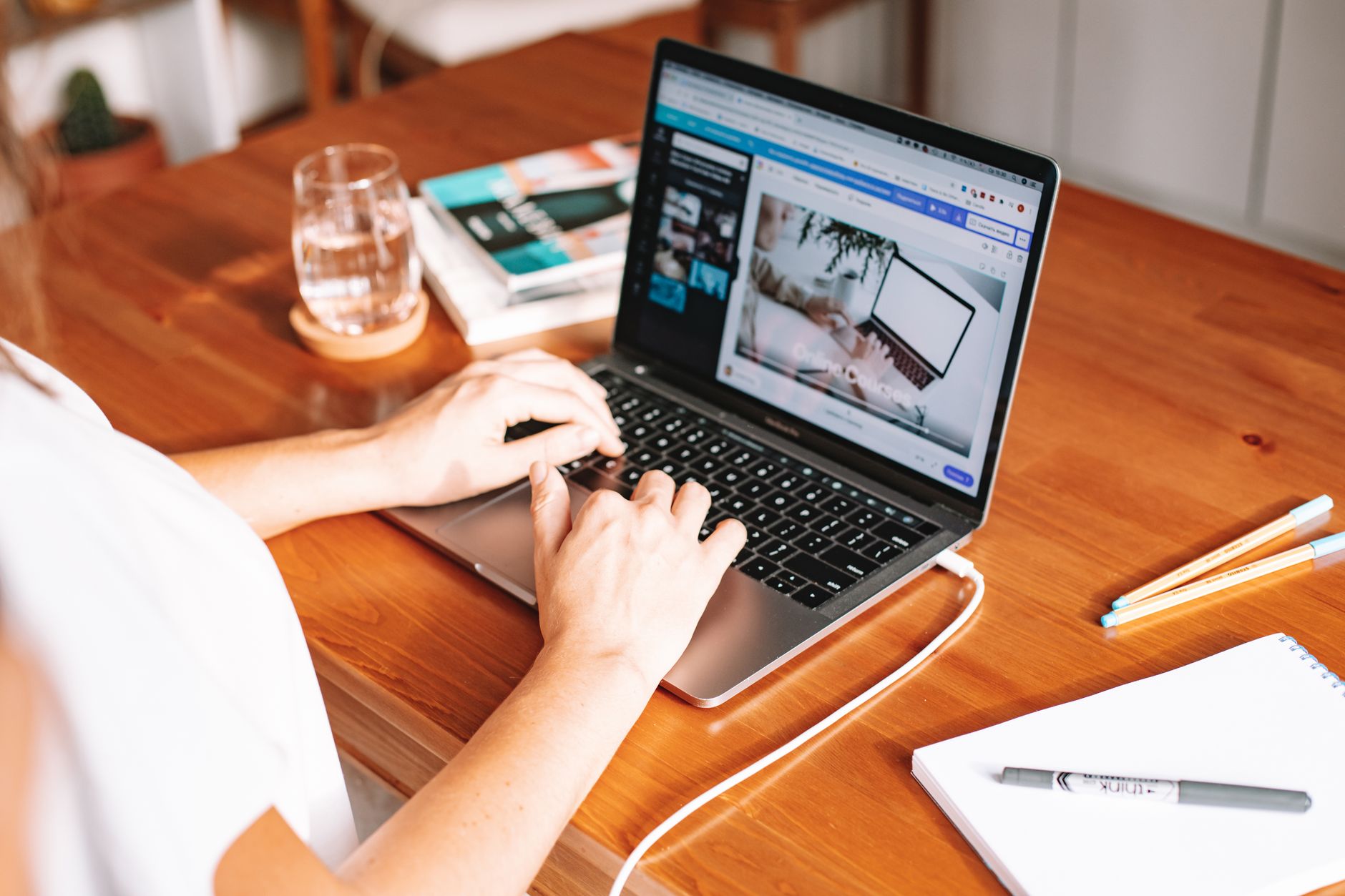 Een persoon werkt vanuit huis aan een laptop aan een houten bureau, met notitieblok en pen.