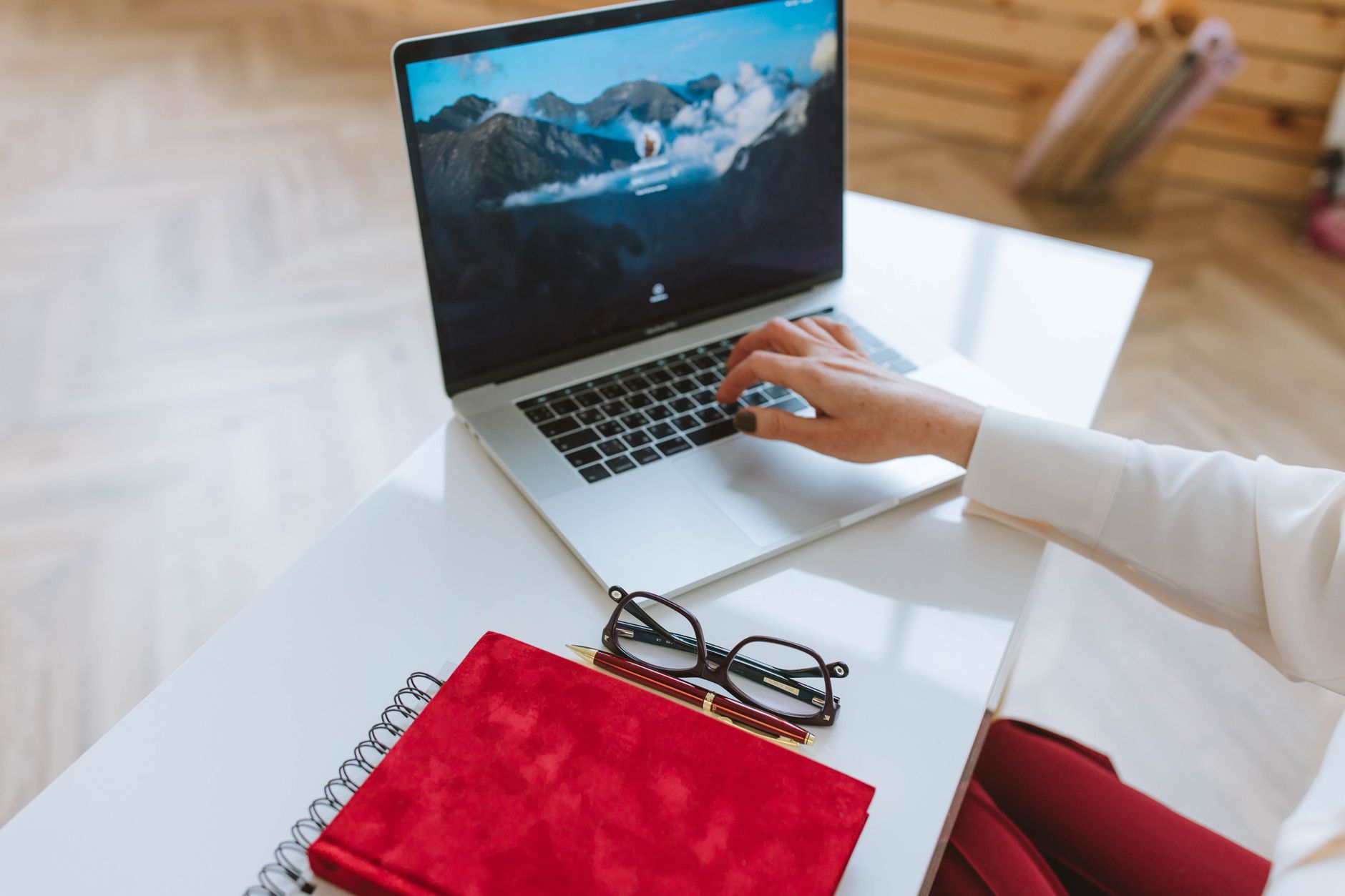 Iemand werkt op een laptop met een rood notitieboek en een bril op een witte tafel.